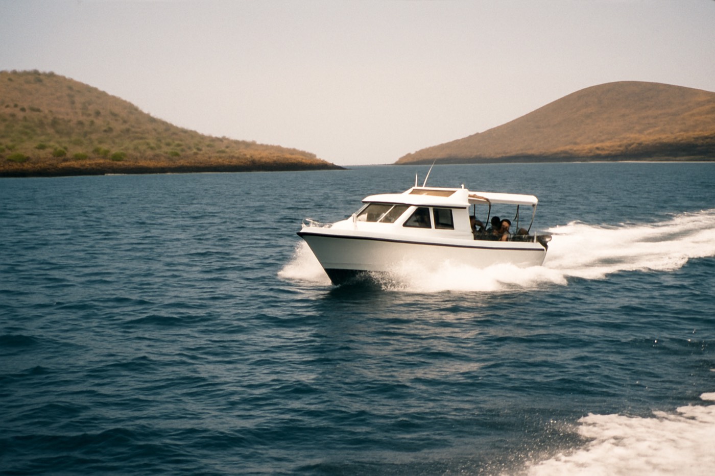 A small white day-tour boat under way in open water between two low volcanic Galapagos islands