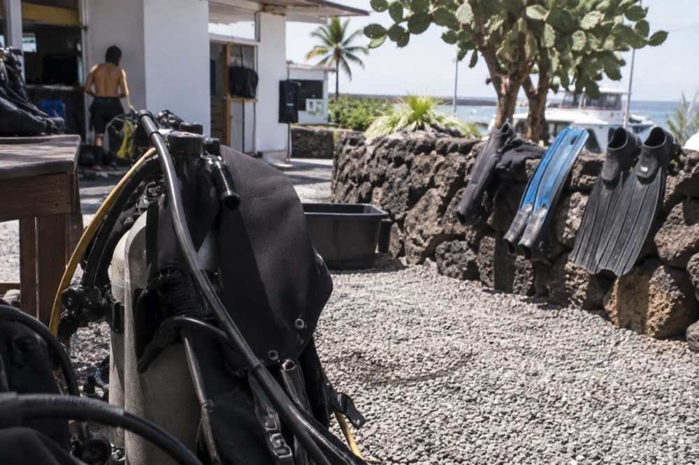Midday glare in a Puerto Ayora dive school courtyard in Ecuador's Galapagos, a trainee's wetsuit half peeled over a dive tank on gravel