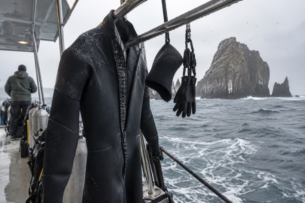 A thick 7mm wetsuit hangs dripping from a rail on a dive deck off Ecuador's Galapagos, September drizzle and cold upwelling chop below