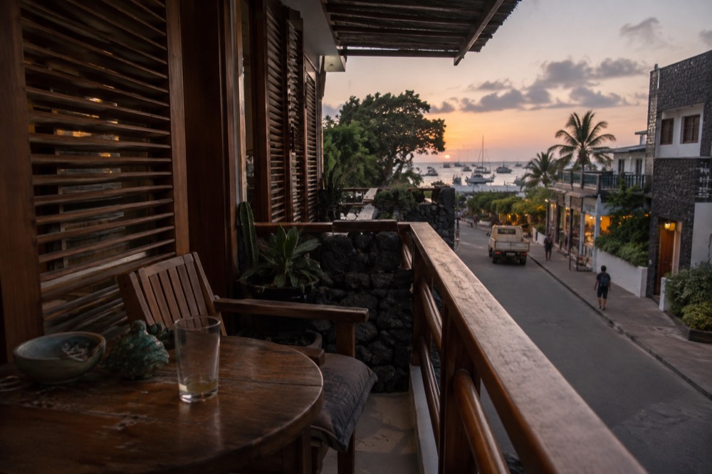 Evening on a guesthouse balcony above a dust-rinsed street in Puerto Ayora, Ecuador, wooden louvred shutters, a half-empty glass on a scuffed table