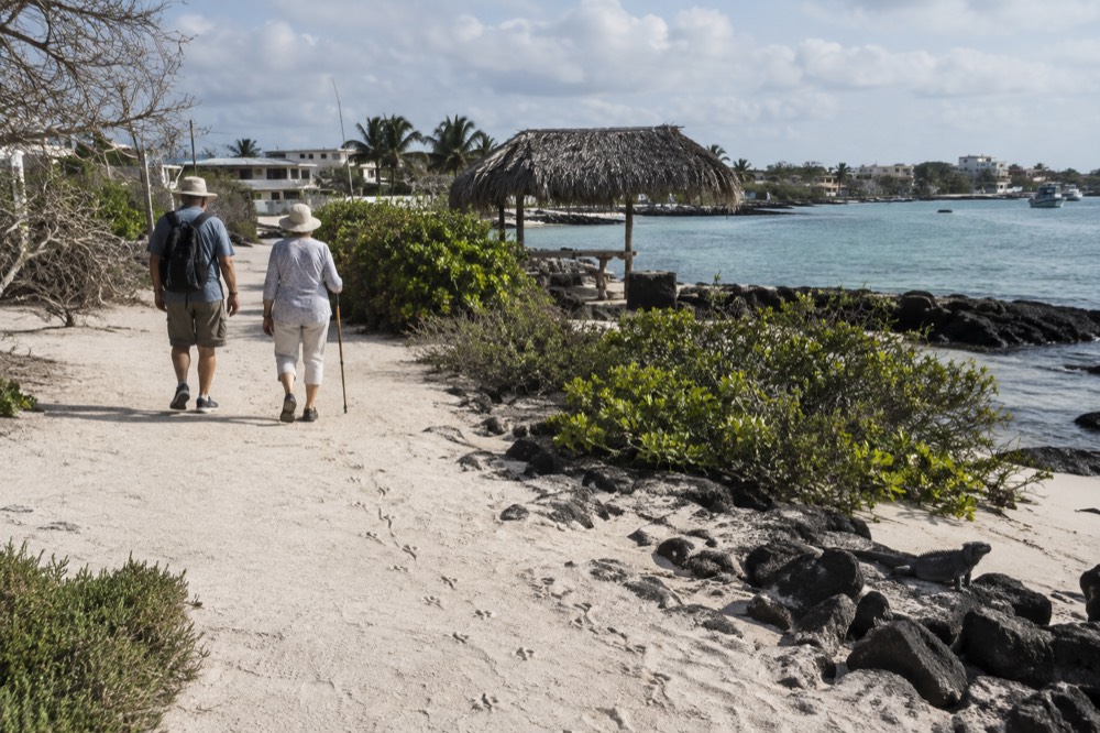 Two older visitors stroll the flat coral-sand path along the Puerto Villamil waterfront, Ecuador, mid-morning sun through salt-bent beach vegetation