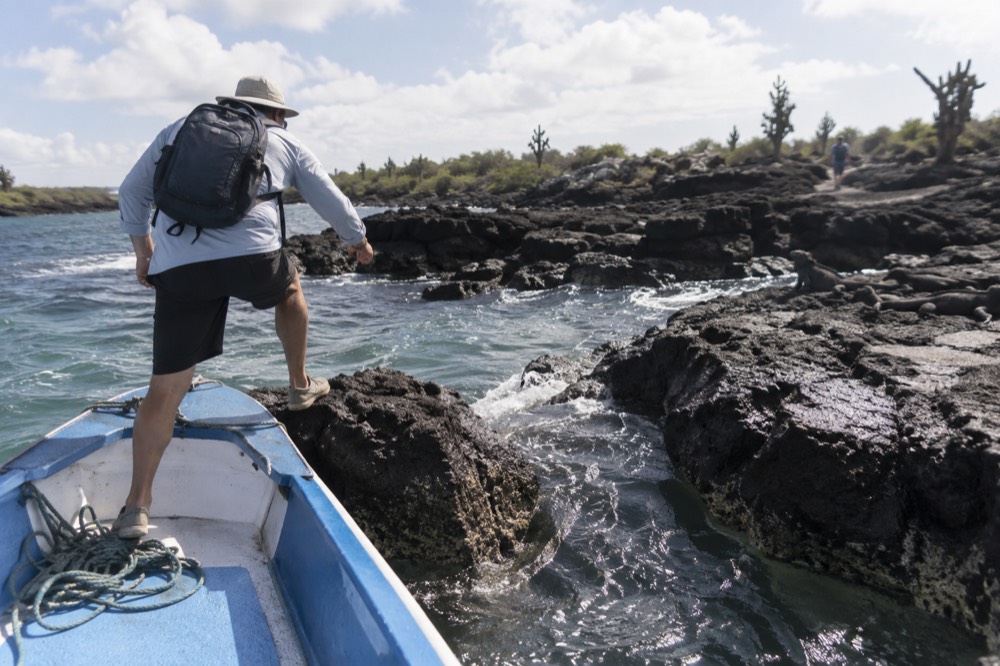 A panga nose ties to a dark lava ledge at Las Tintoreras, Galapagos Islands, Ecuador, a traveller stepping across the closing gap onto wet rock