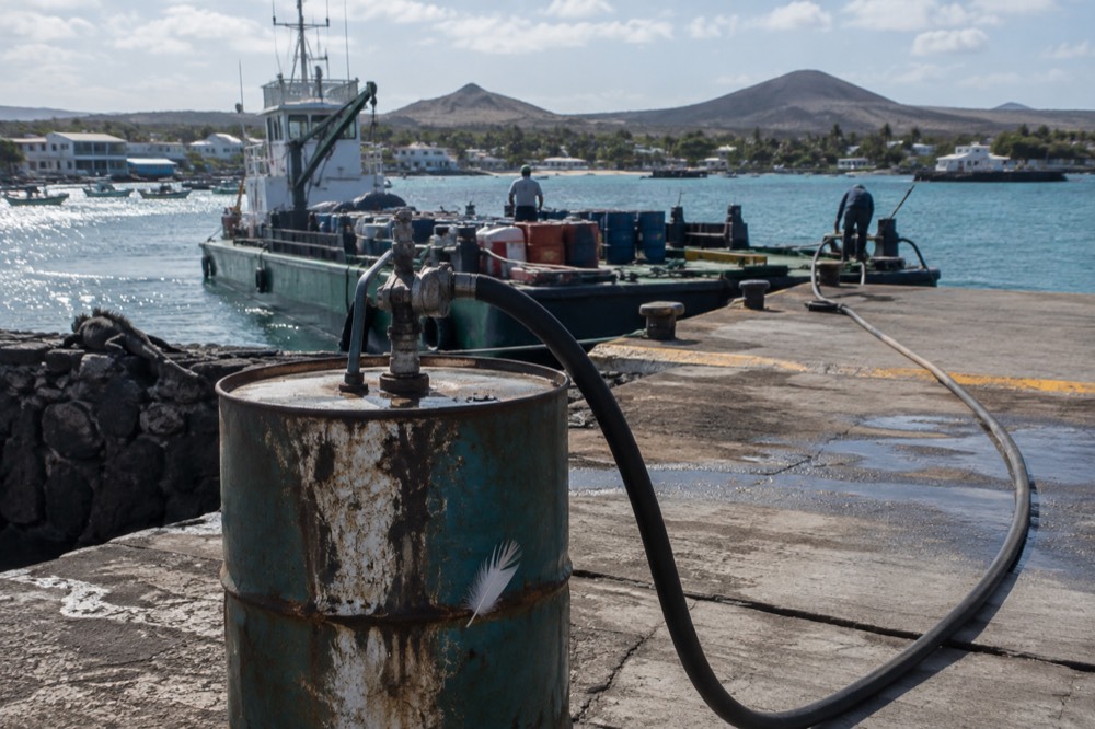 Hot midday at a fuel depot dock in Puerto Ayora, Galapagos, a supply barge and volcanic ridgeline soft beyond, glare off the water