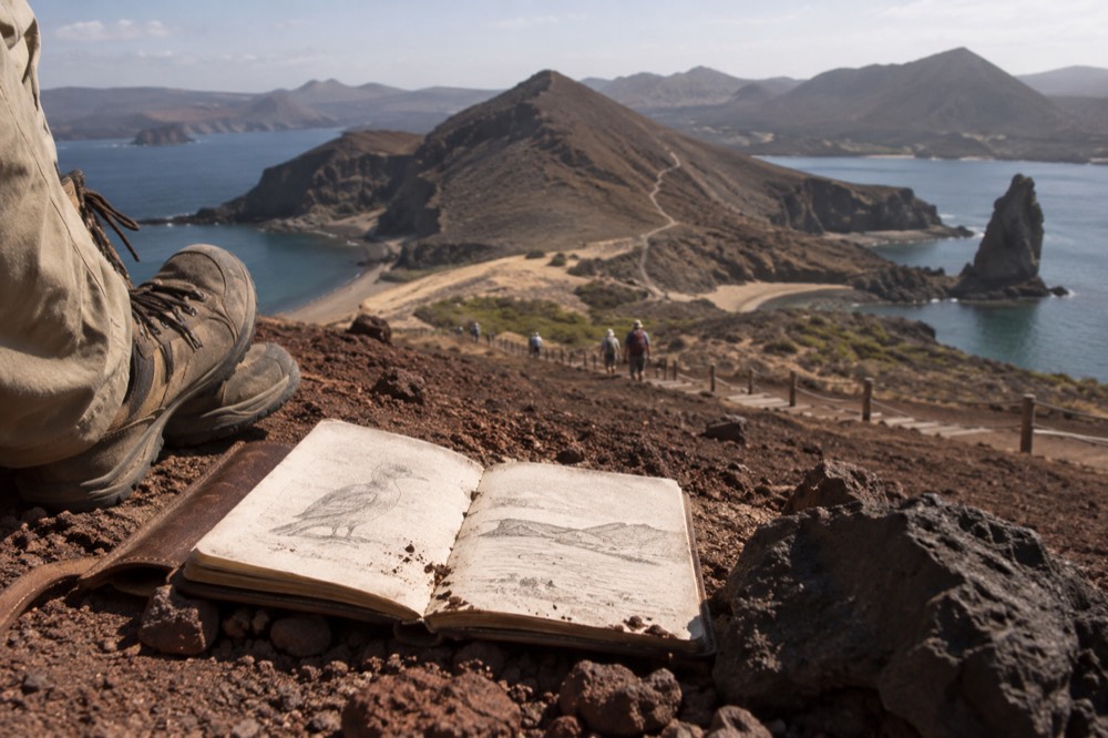 A naturalist guide's worn leather field journal and scuffed hiking boots on the rust-red volcanic gravel of Bartolomé Island, Galapagos Islands, Ecuador