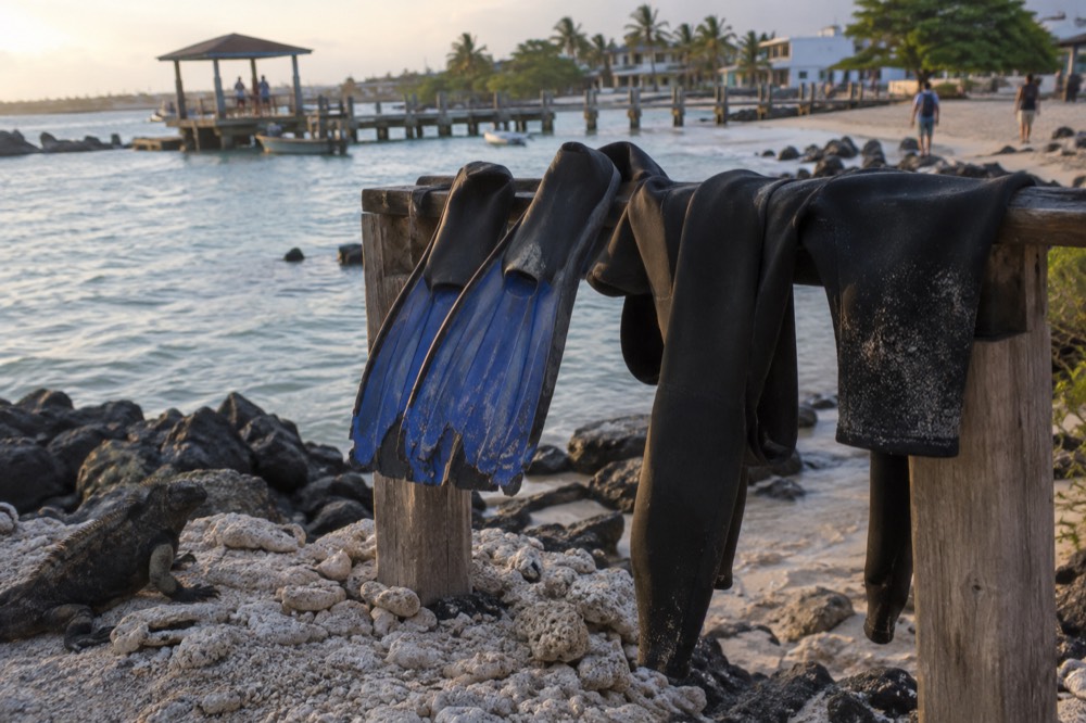 Torn snorkel fins and a damp wetsuit drying on a wooden rail at Villamil's sandy pier in the Galapagos, Ecuador, warm late-afternoon light